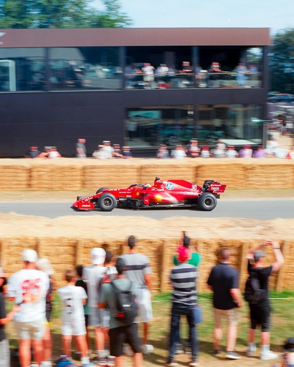 A red race car speeds past a crowd of spectators and a building with large windows.