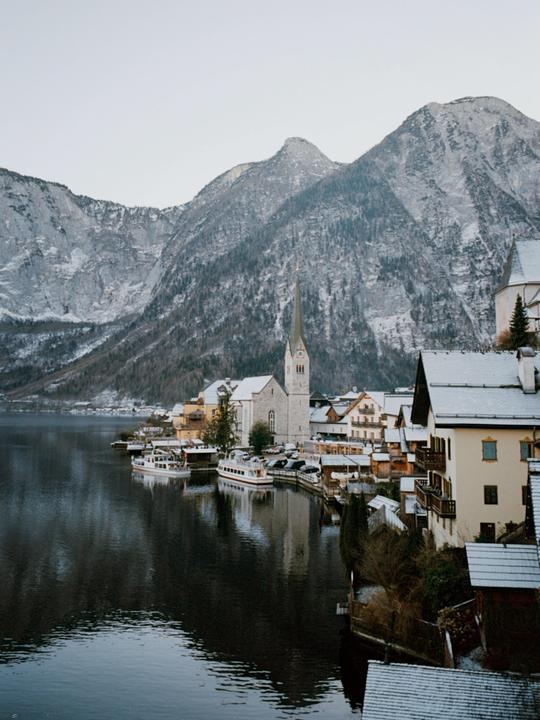 A town with a prominent church steeple is nestled beside a lake, surrounded by snow-dusted mountains.
