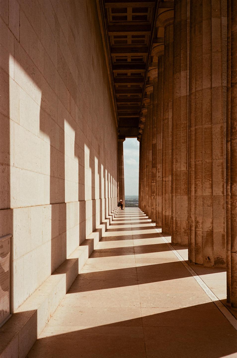 A person sits at the end of a long hallway lined with columns and shadows.