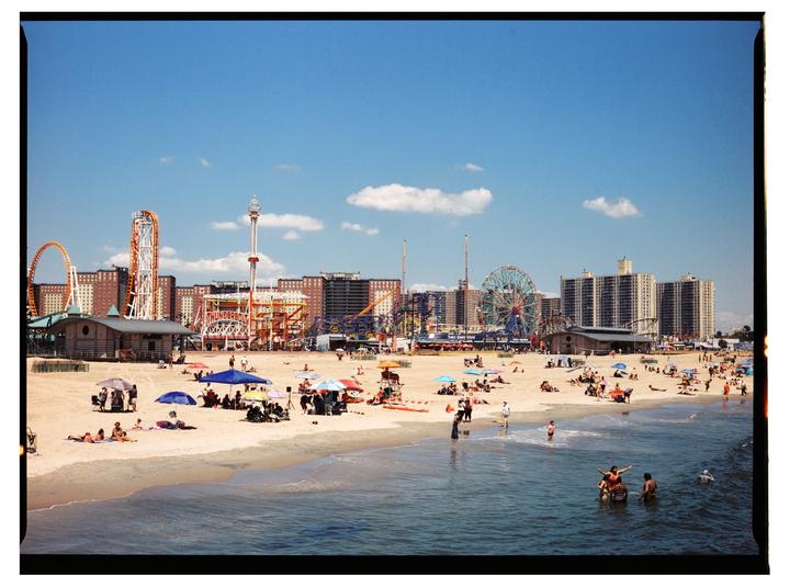 A wide shot shows a crowded beach with an amusement park and buildings in the background under a blue sky.