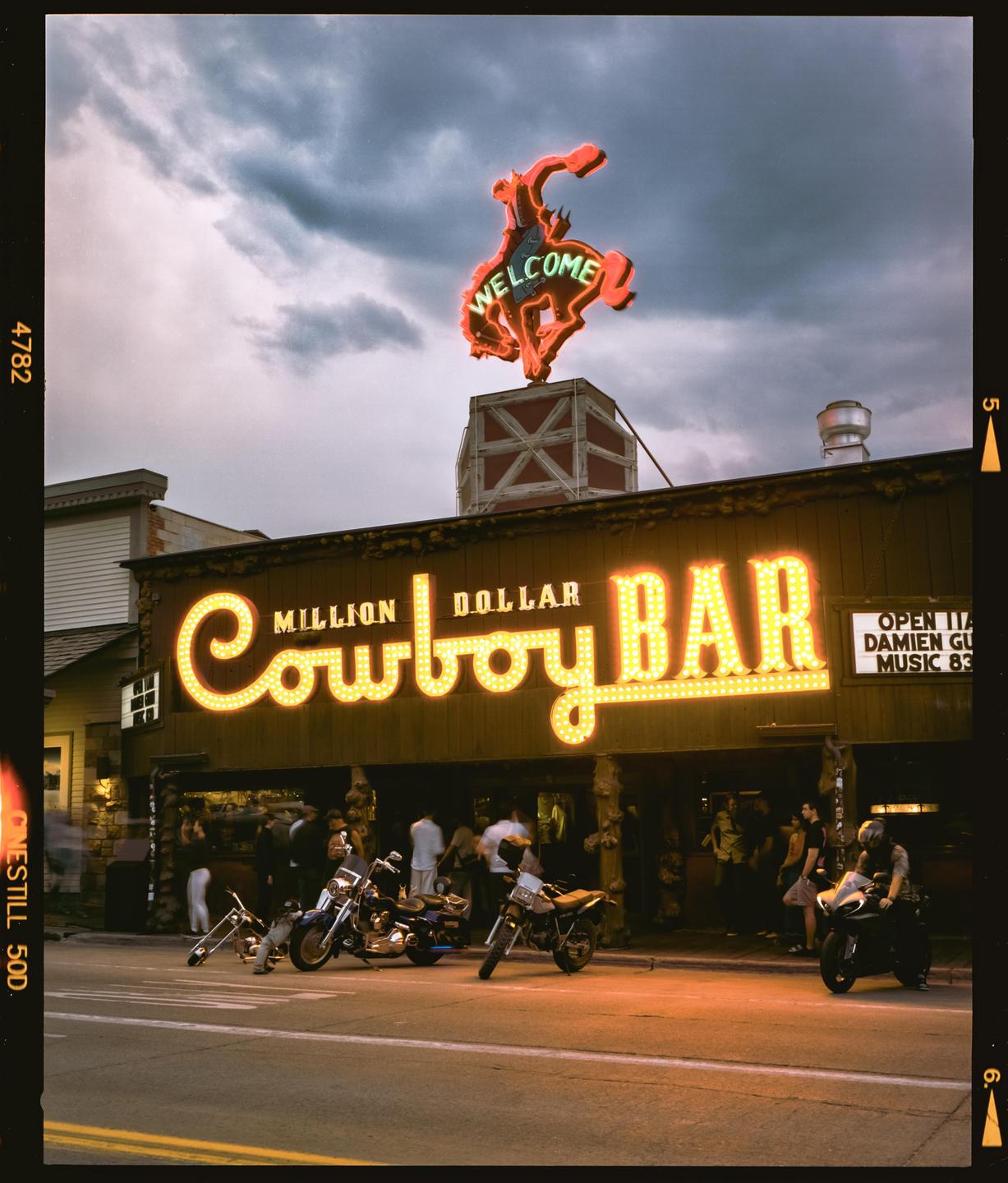 A neon cowboy sign and a bright "Million Dollar Cowboy Bar" sign illuminate a street with motorcycles parked in front of a bar.