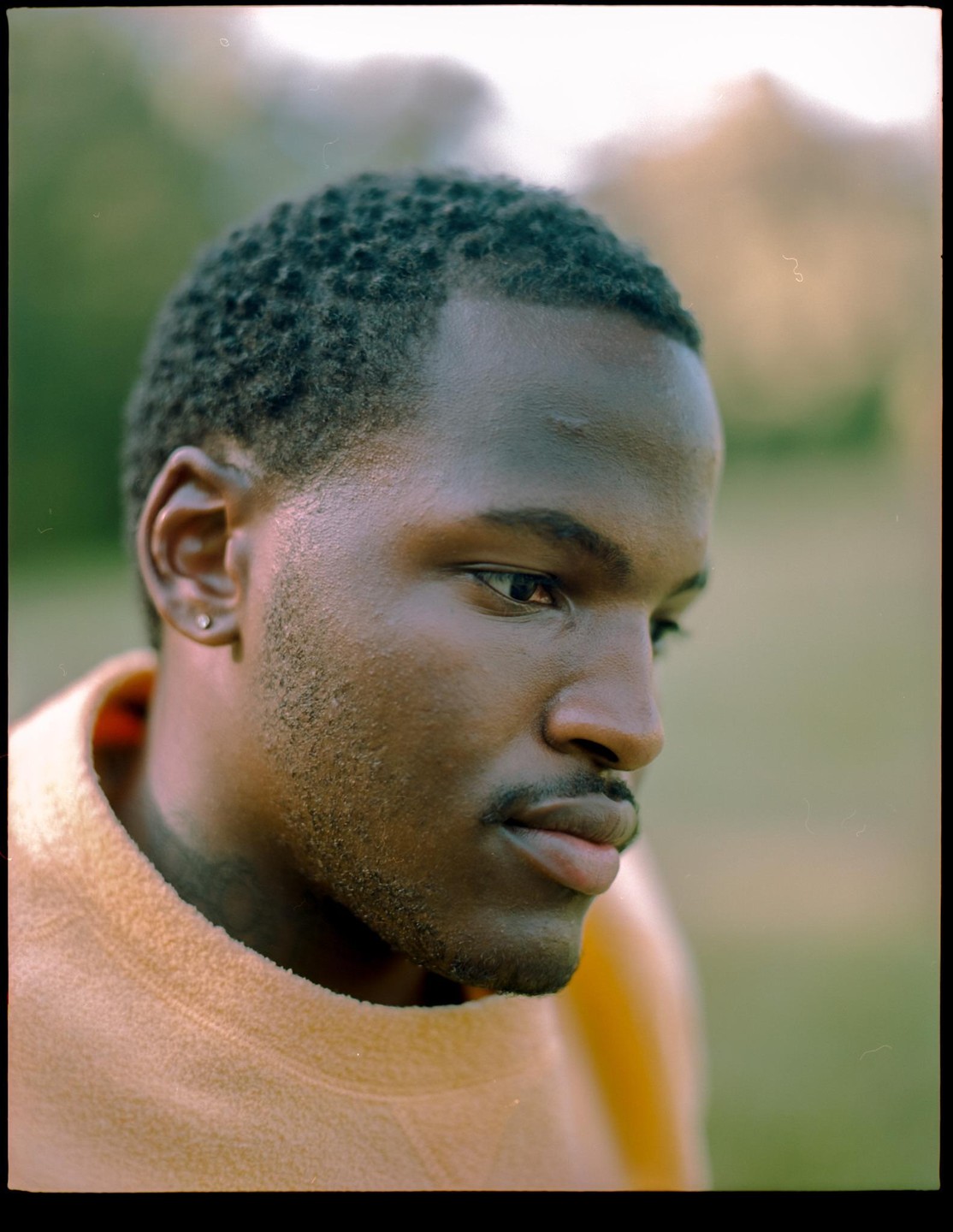 A man with short hair and a mustache looks to the right of the frame, wearing an orange sweater.