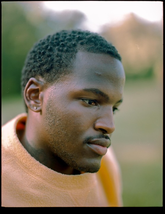 A man with short hair and a mustache looks to the right of the frame, wearing an orange sweater.