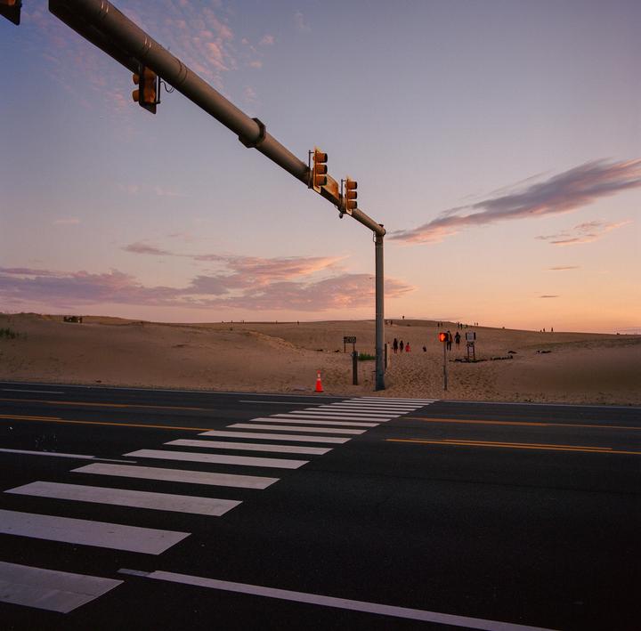 A crosswalk leads to a sandy dune landscape with traffic lights overhead at sunset.