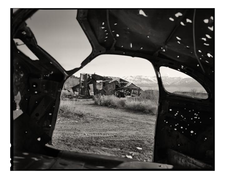 A dilapidated wooden building and snowy mountains are visible through the frame of a wrecked vehicle.