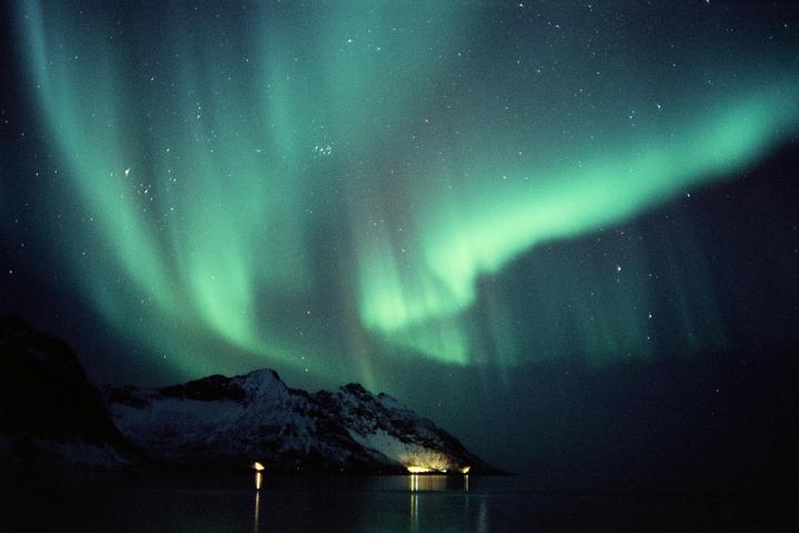 The aurora borealis glows green above a snowy mountain and a body of water with lights reflecting on its surface.