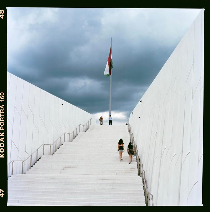 A Hungarian flag flies above a long, wide, white staircase with people walking up it under a cloudy sky.