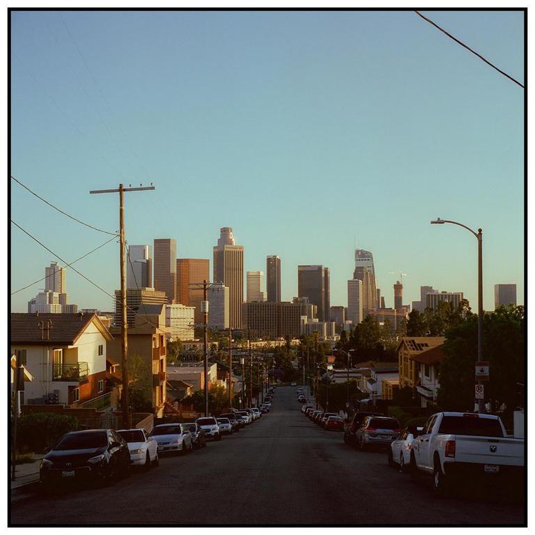 A street lined with parked cars leads to a distant city skyline under a clear sky.
