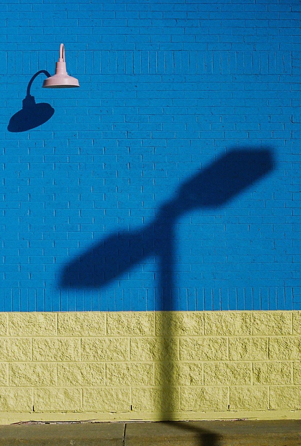 A pink light fixture casts a shadow on a blue and yellow brick wall.