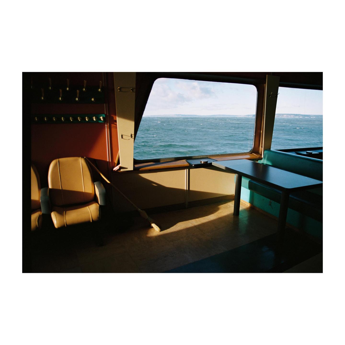 Sunlight streams through the window of a ferry, illuminating a broom on the floor and the ocean outside.