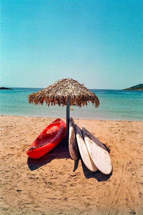 A red kayak and three surfboards rest on a sandy beach under a straw umbrella, with the Aegean Sea and distant land visible in the background.