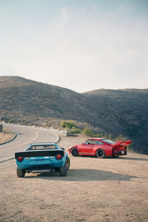 Two sports cars, one blue and one red, are parked on a dirt pull-off next to a winding road on a hillside.