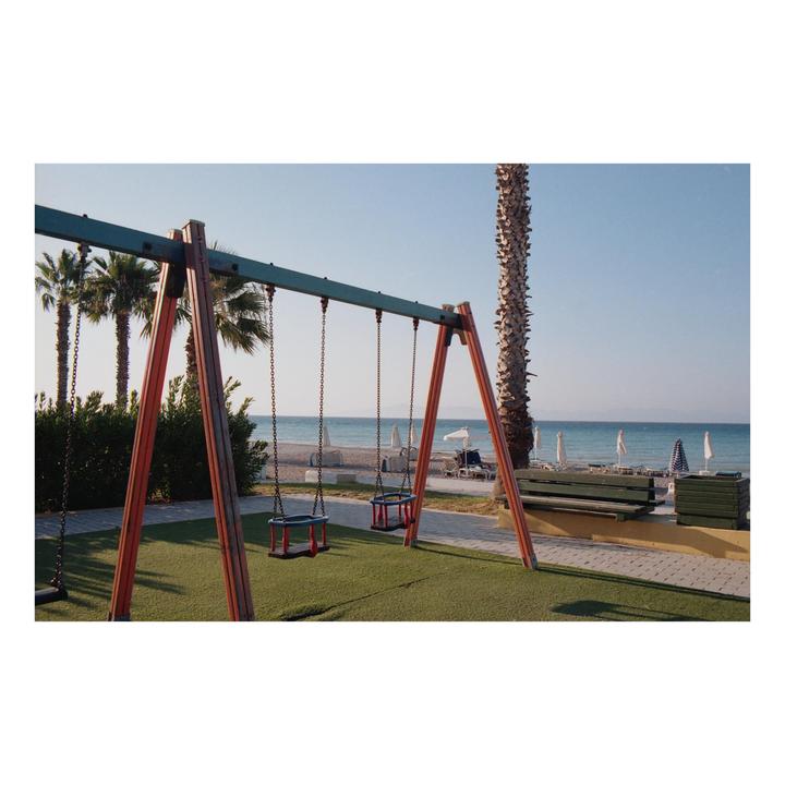 A swing set sits on artificial grass next to a beach with palm trees and lounge chairs.