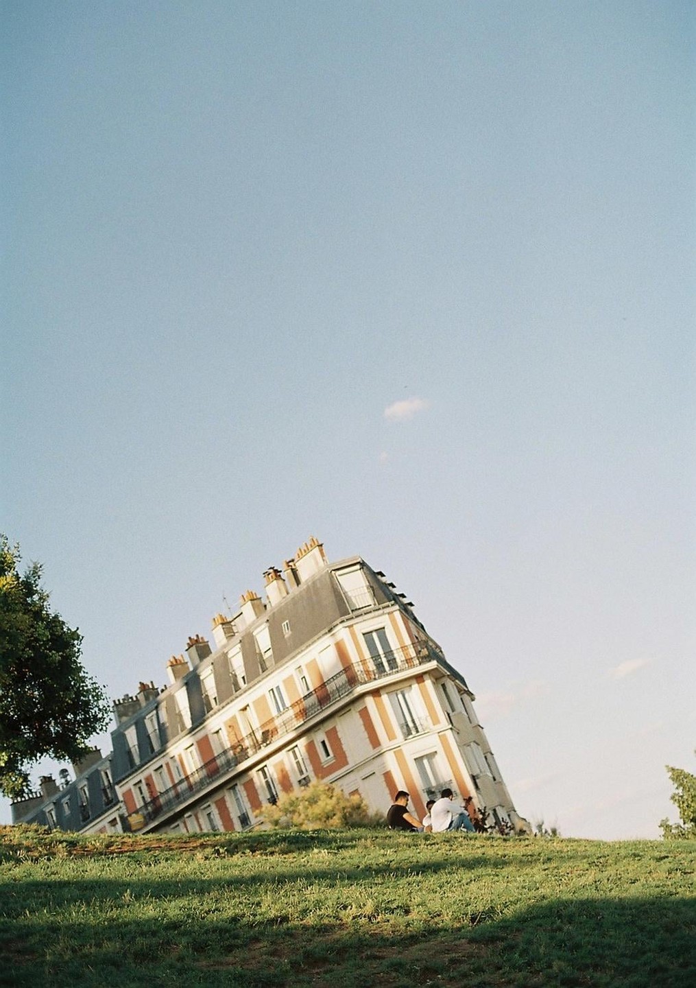 A building appears to be sinking into a grassy hill with two people sitting on the grass in the foreground.