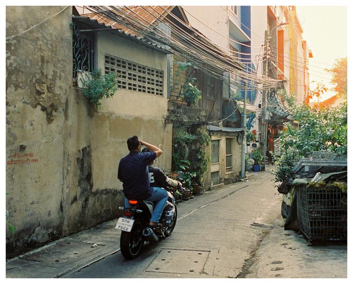 A man rides a motorcycle down a narrow street lined with buildings and lush greenery.