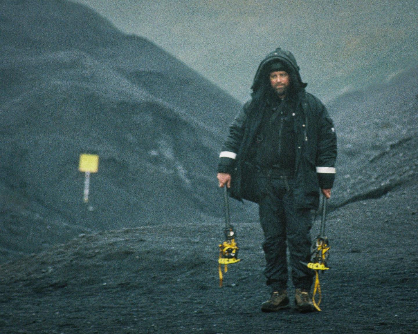 A man in a hooded jacket and boots stands on a dark, rocky landscape holding two ice axes.