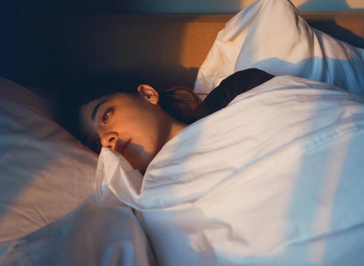 A woman lies in bed, partially covered by a white duvet, with warm light illuminating her face.