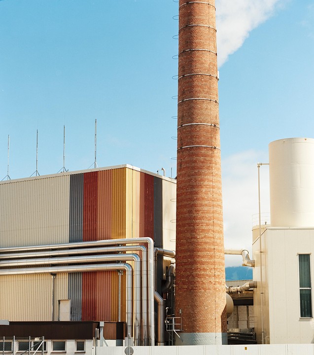 A tall brick chimney stands next to an industrial building with colorful striped panels and silver pipes under a clear blue sky.