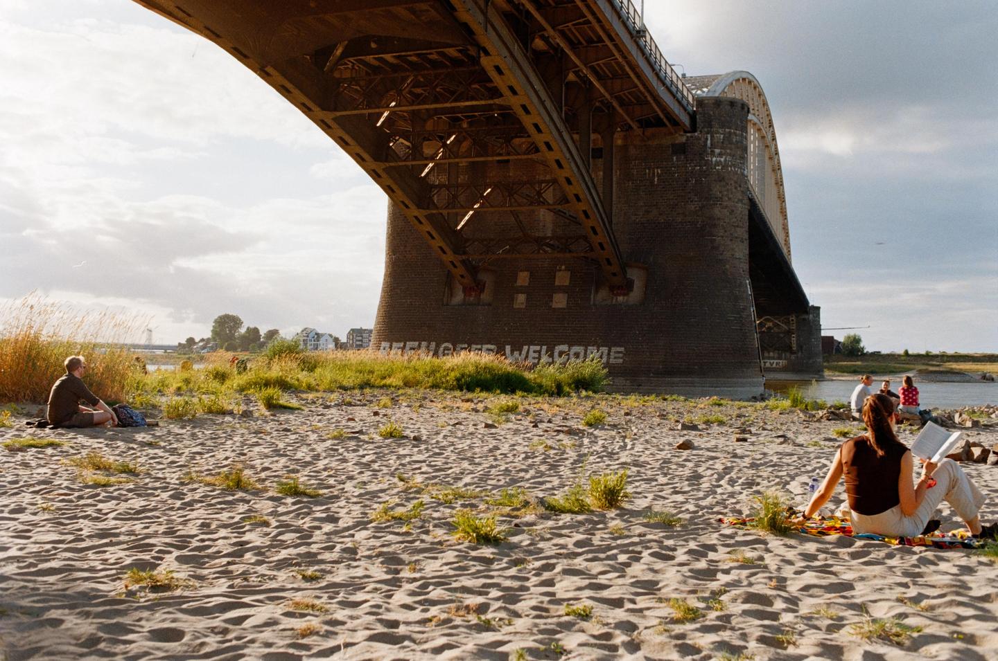 A woman reads a book on a sandy riverbank under a large bridge, with other people and a town visible in the distance.