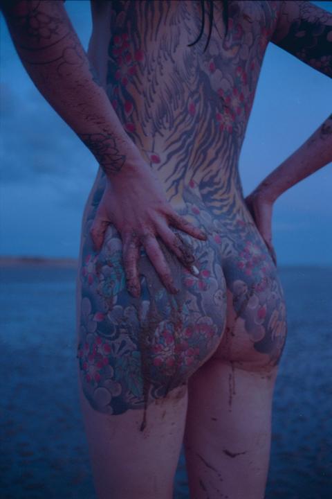 A person with full body tattoos stands with their back to the camera, covered in mud, by the sea at dusk.