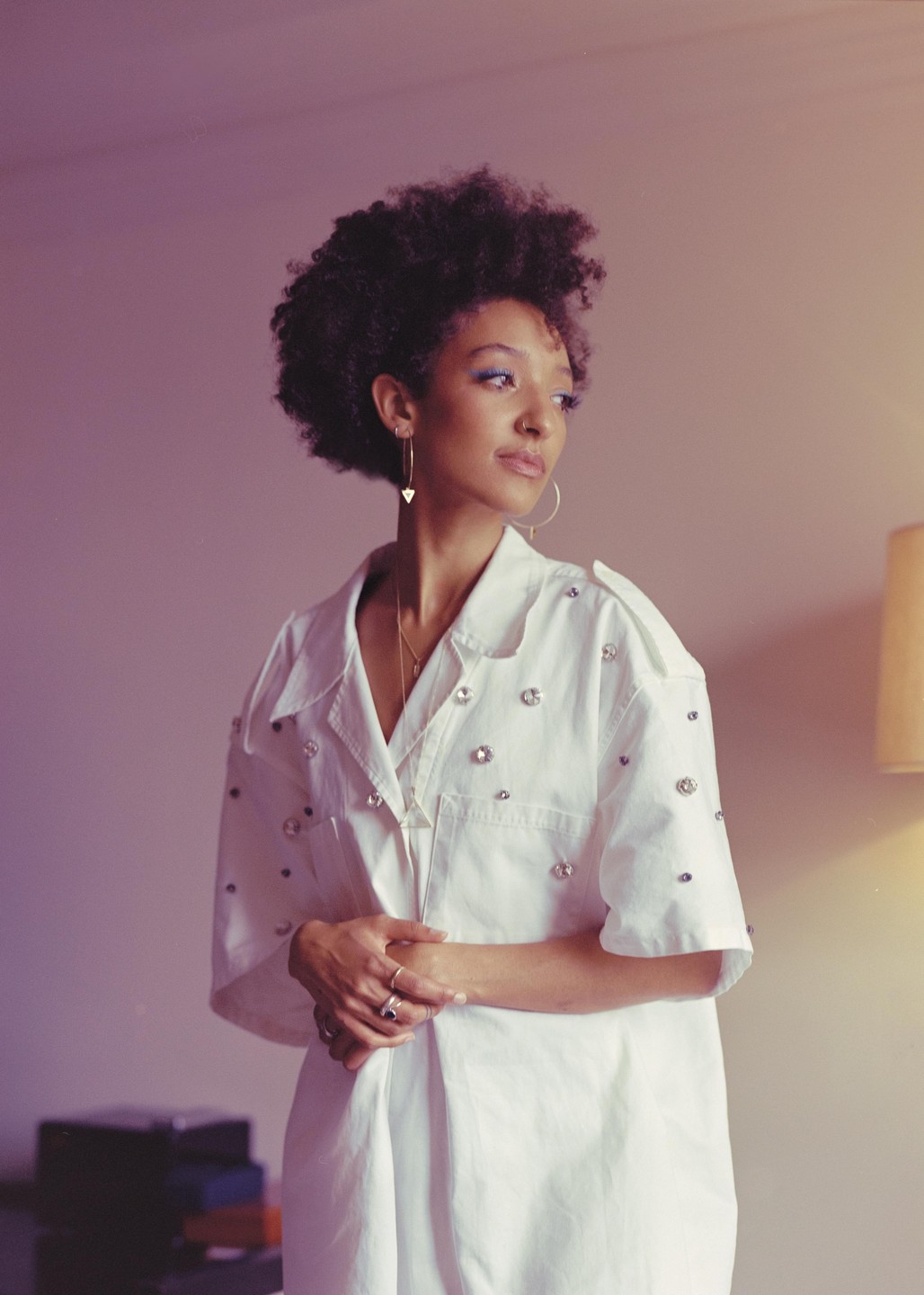 A woman with curly hair and a white shirt with embellishments looks to the side.