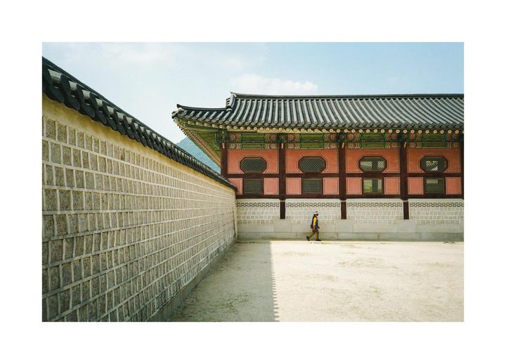 A person walks along a dirt path next to a stone wall and a traditional Korean building with a tiled roof.