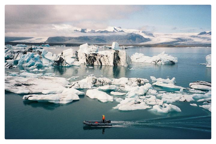 A person in a small boat navigates through a glacial lagoon filled with icebergs, with snow-capped mountains in the background.