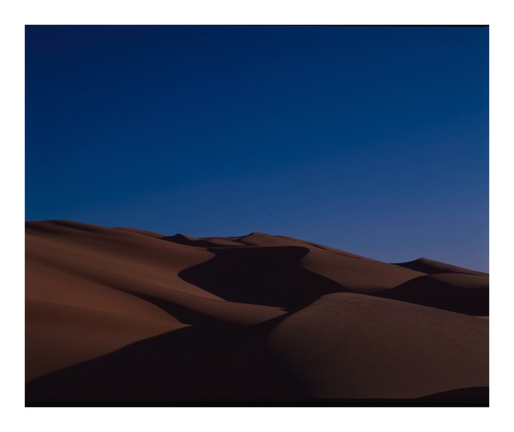 A desert landscape shows sand dunes under a clear blue sky.