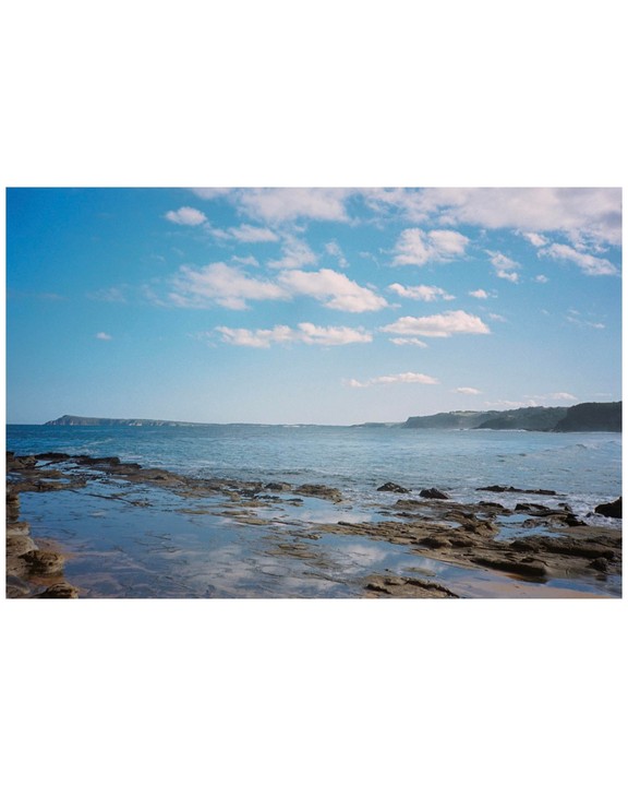 A rocky coastline with tide pools reflects the sky under a blue sky with scattered clouds.