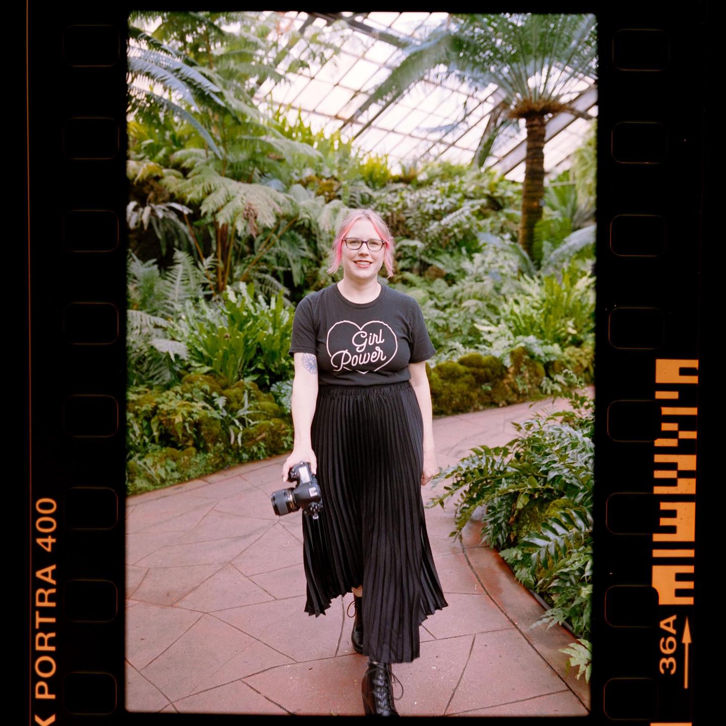A woman with pink hair and glasses smiles at the camera while holding a camera in a lush greenhouse.