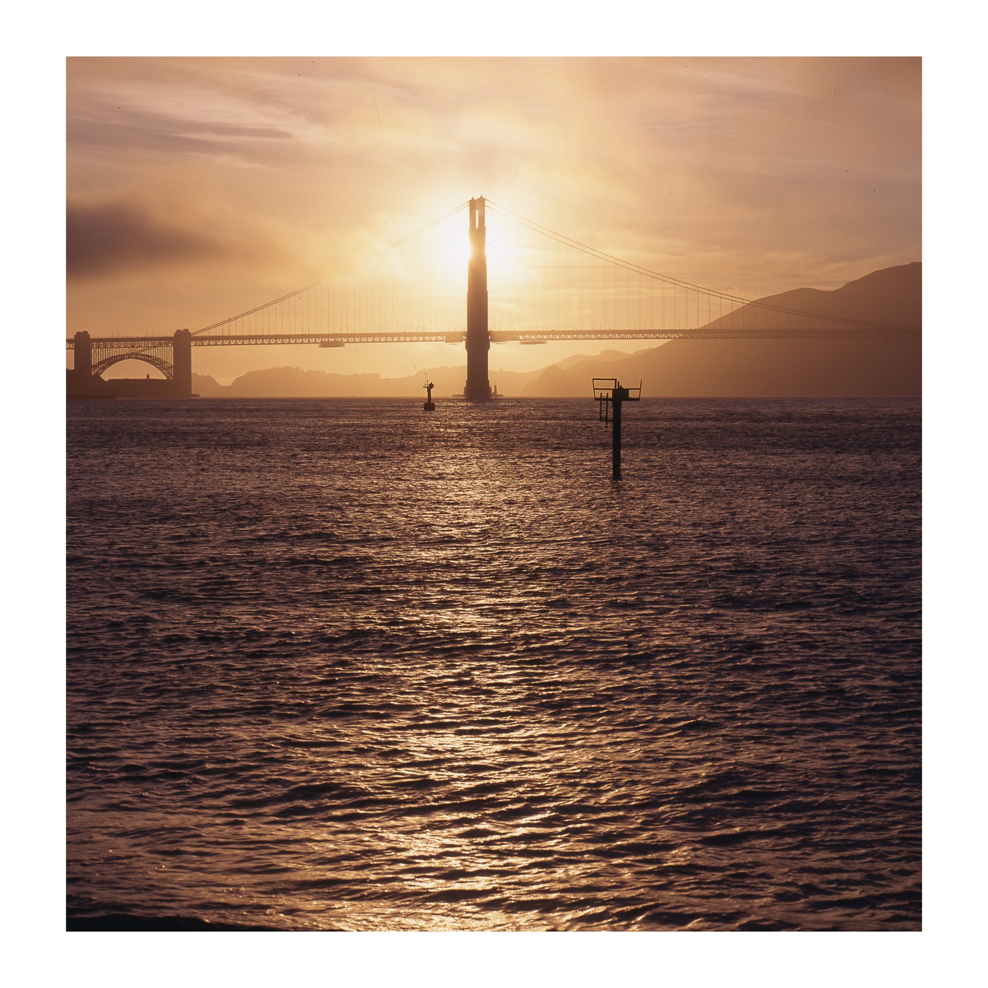 The Golden Gate Bridge is silhouetted against a bright sunset over the water.