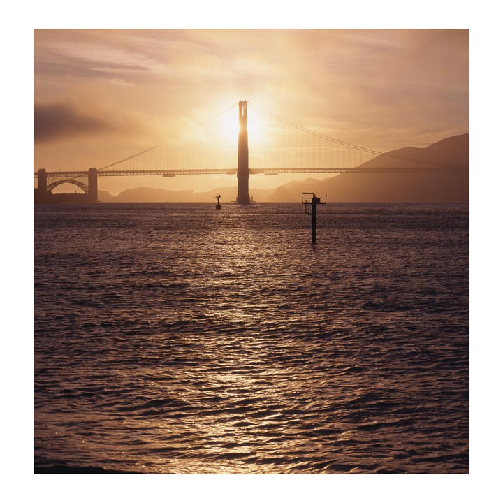 The Golden Gate Bridge is silhouetted against a bright sunset over the water.