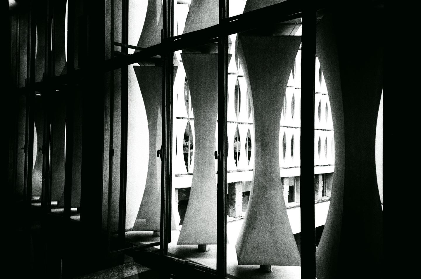 A dark interior looks out through a window with vertical concrete fins at a building with oval windows.