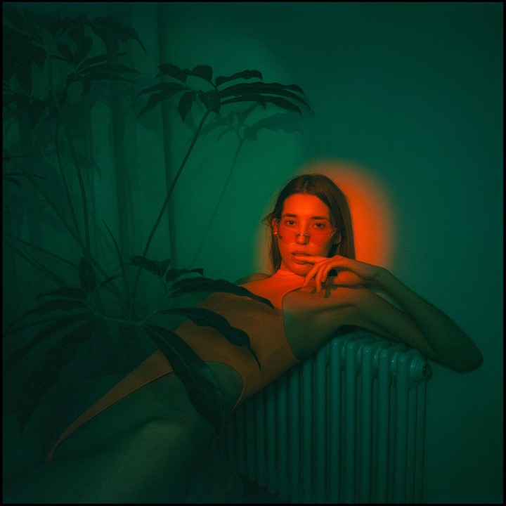 A woman in a swimsuit and sunglasses sits next to a plant and a radiator, illuminated by green and orange light.