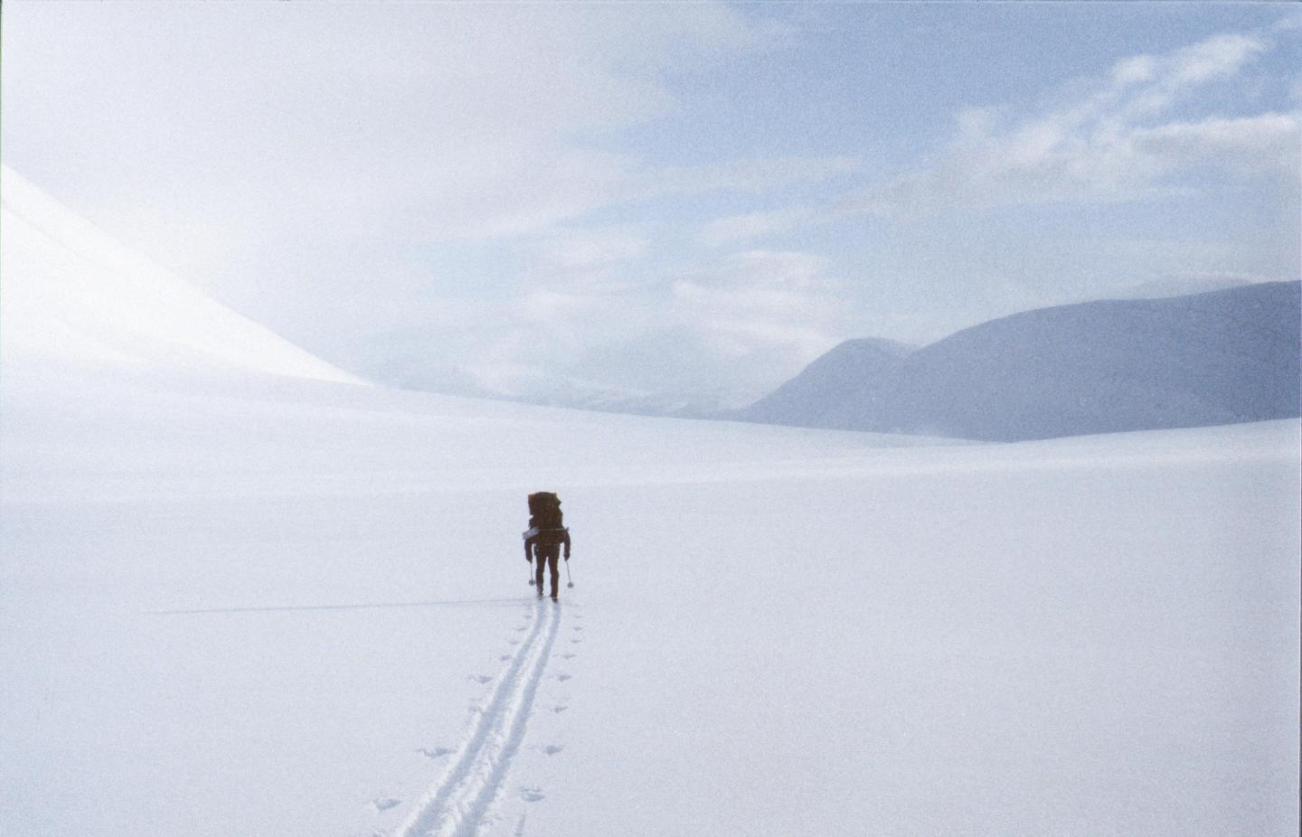 A person with a large backpack skis across a vast, snow-covered landscape with mountains in the distance.