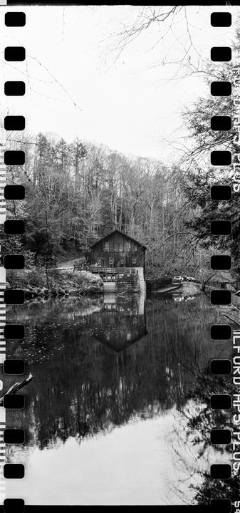A wooden mill building stands beside a reflective body of water, surrounded by a forest of bare trees.