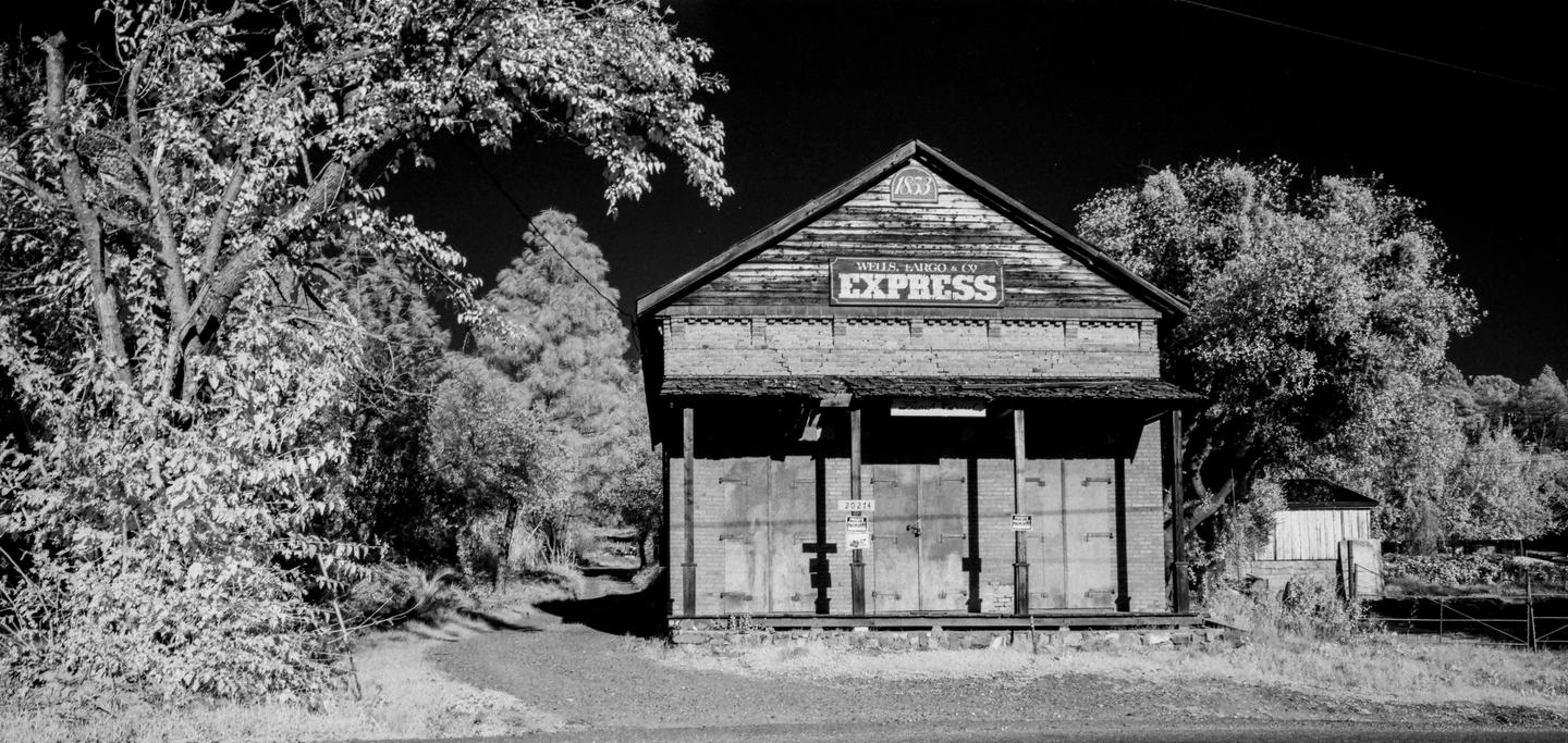 An old wooden building with a "Wells, Fargo & Co Express" sign on its facade stands next to a dirt path and trees.