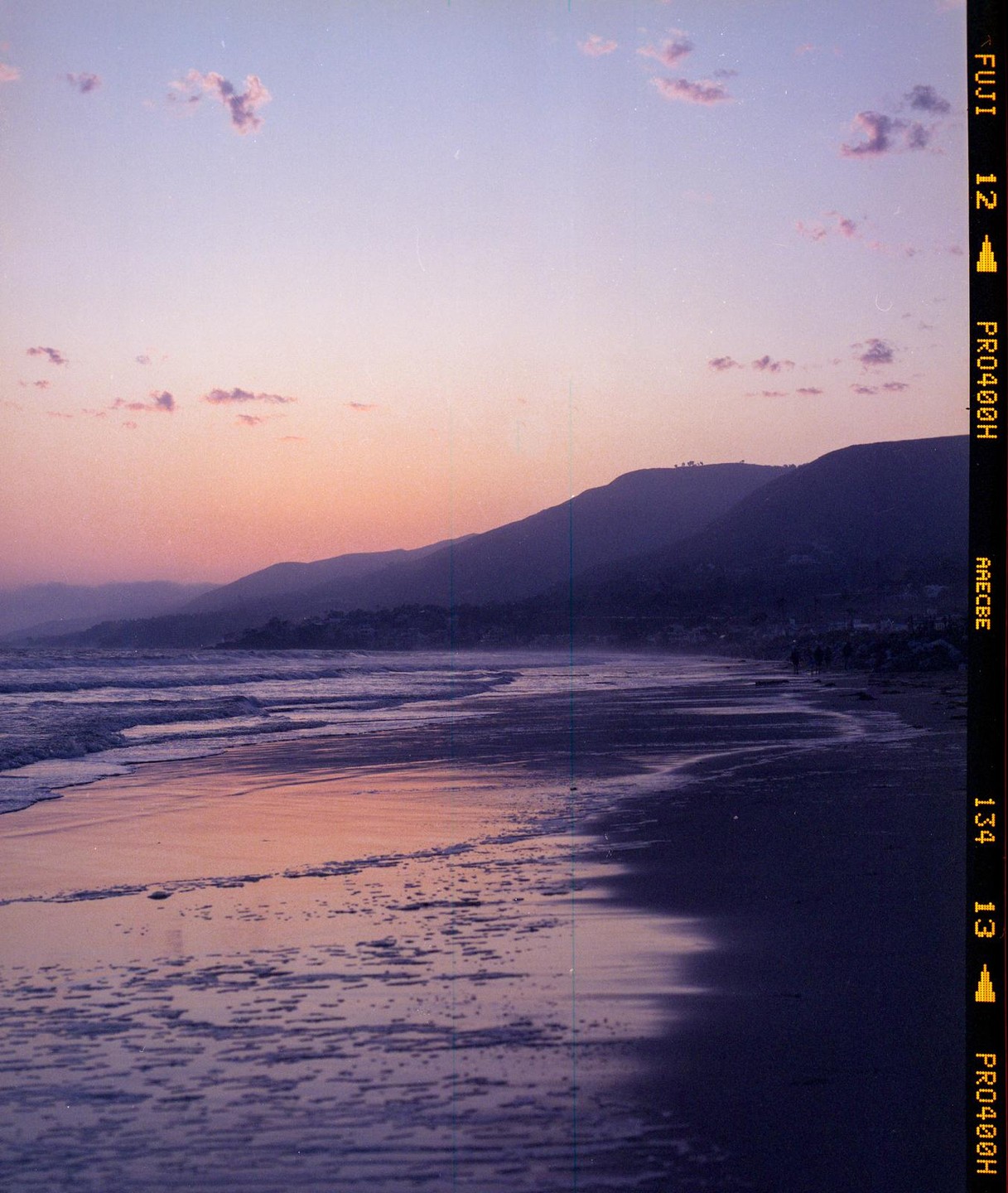 Waves wash onto a beach at sunset with mountains in the background.