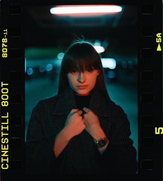 A woman with bangs and long hair wears a coat and watch, illuminated by red and blue light at night.