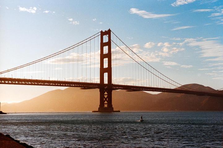 A person on a surfboard is in the water in front of a large suspension bridge with mountains in the background.