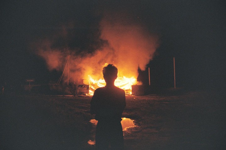 A person stands with their back to the camera, silhouetted against a large fire burning a structure at night.