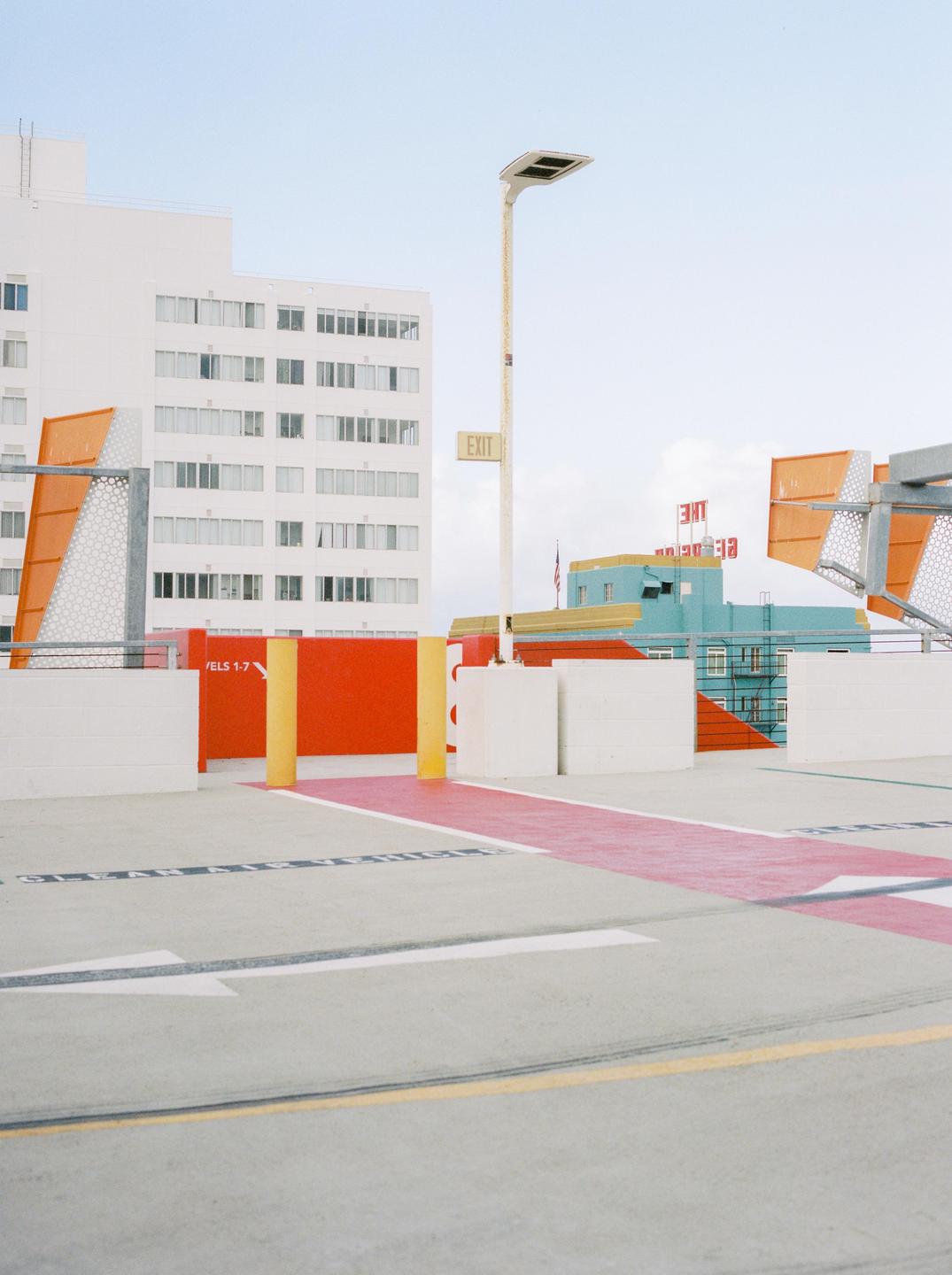 A parking garage with a red path and yellow poles leads to a white building and a turquoise building with a red sign.