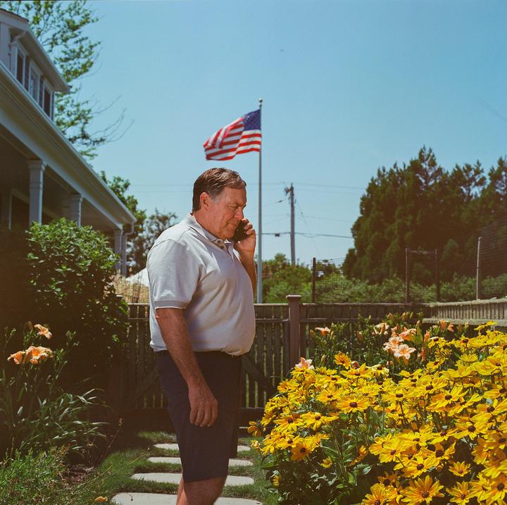 A man talks on a phone in a garden with a house and an American flag in the background.
