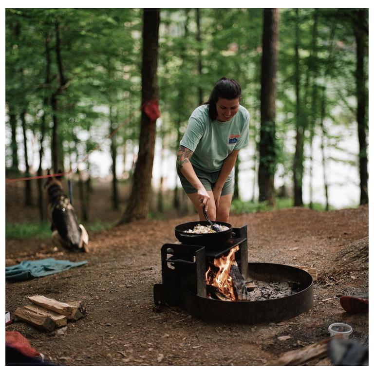 A woman cooks food in a pan over a campfire in a forest with a dog in the background.