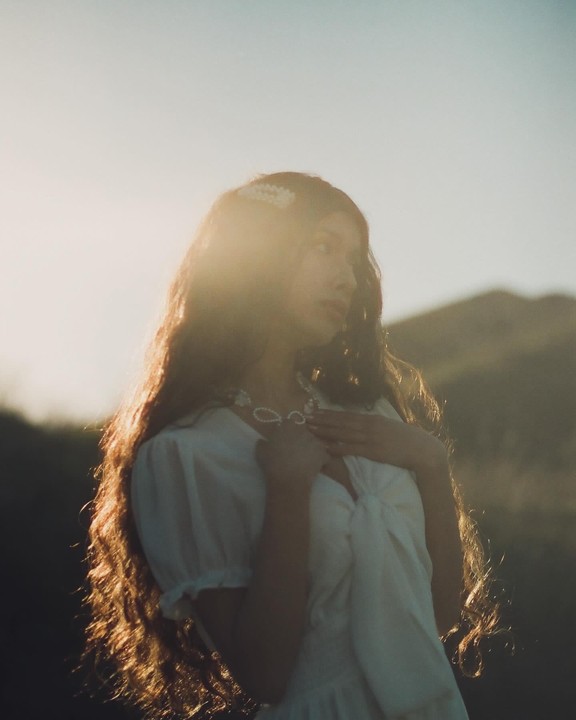 A woman with long, wavy hair and a white dress stands outdoors with a mountain in the background.