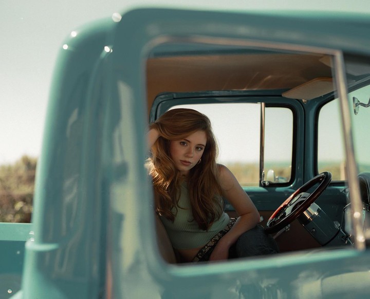 A woman with long hair sits in the driver's seat of a light blue vintage truck.