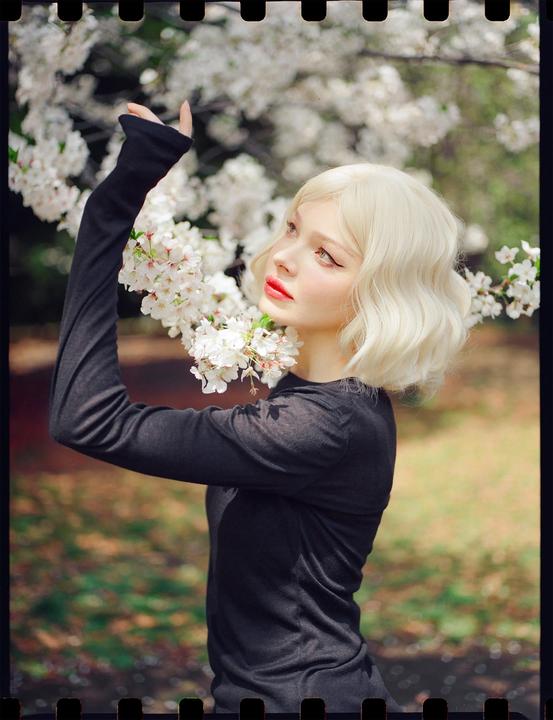 A woman with blonde hair and red lipstick is looking up at white blossoms.
