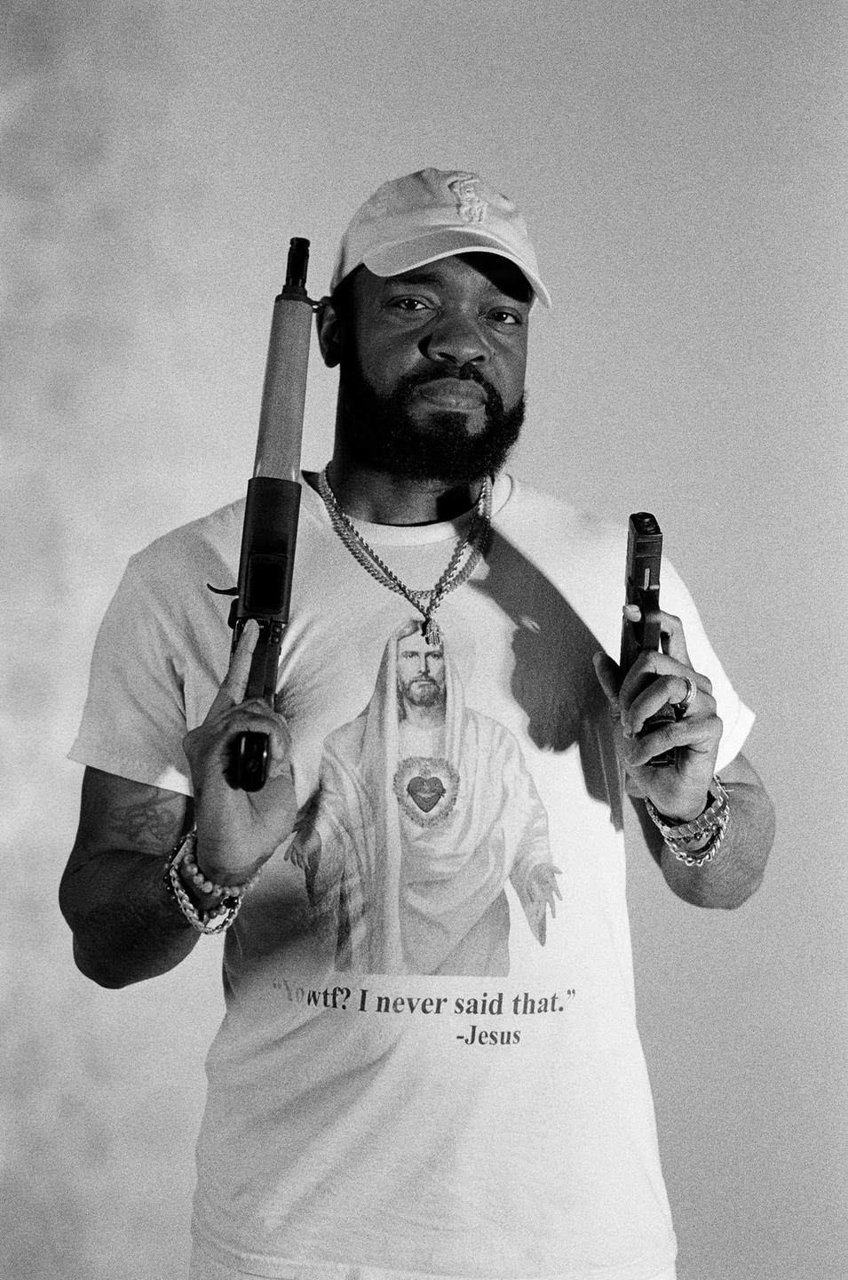 A man wearing a cap and a t-shirt with a graphic of Jesus holds a rifle and a handgun.