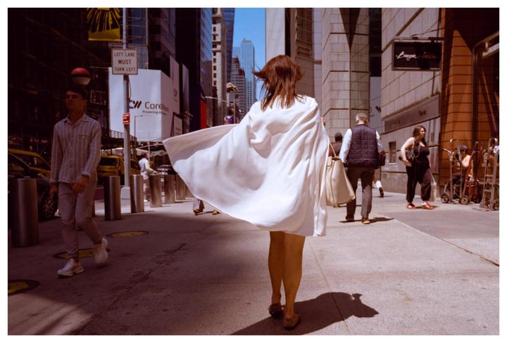 A woman in a white dress walks away from the camera on a city street with other pedestrians and tall buildings.