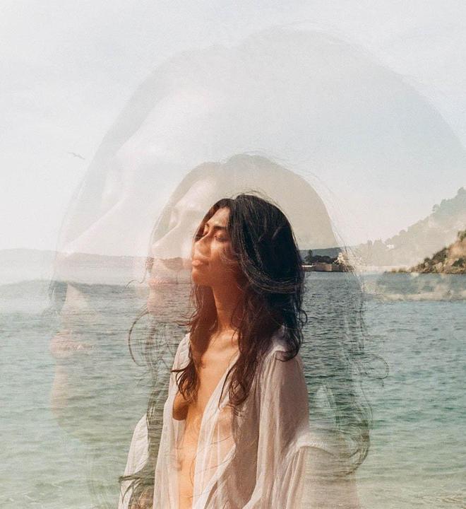 A woman with long dark hair stands in front of a body of water with a hazy sky and a faint outline of a face in the background.